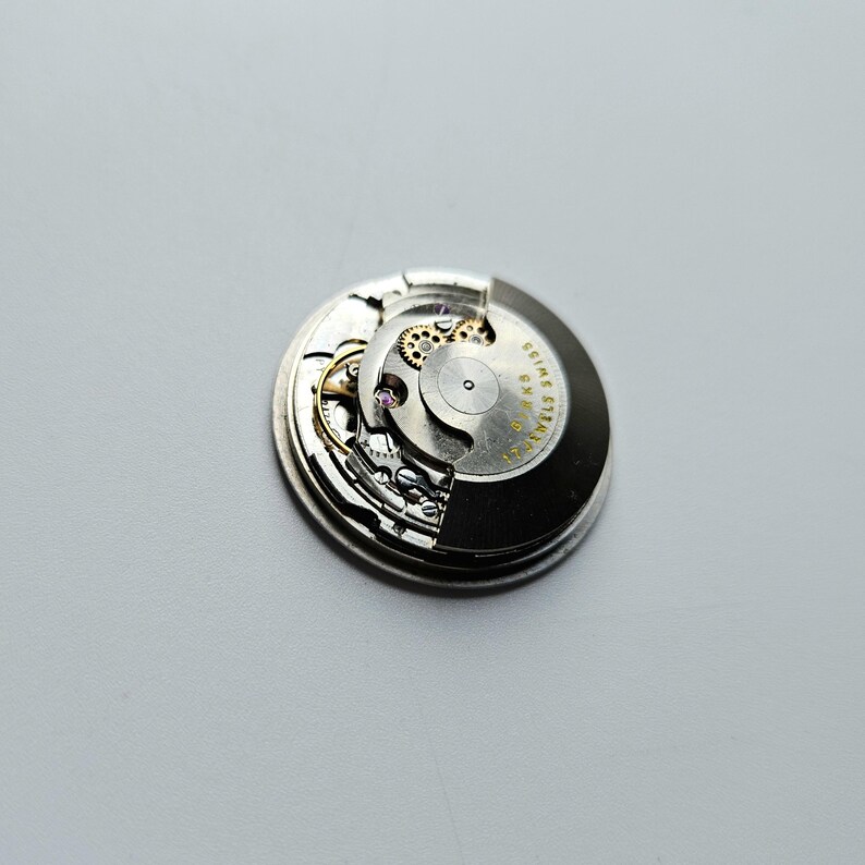 K&ouml;nnte beinhalten: Nahaufnahme eines Uhrwerks mit komplizierten Zahnr&auml;dern und Komponenten. Der silberfarbene Mechanismus weist kleine Schrauben, eine Unruh und den Text "SWISS" und "17 JEWELS" auf. Das Uhrwerk ist vor einem hellgrauen Hintergrund platziert.