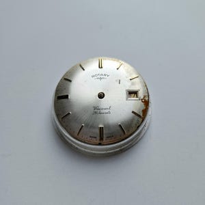 Pode incluir: Um mostrador de relógio prateado com a marca "Rotary" e o texto "Viscount 21 Jewels". O mostrador do relógio tem uma janela de data exibindo o número 7. O mostrador tem um acabamento em metal escovado com marcadores de hora pretos.