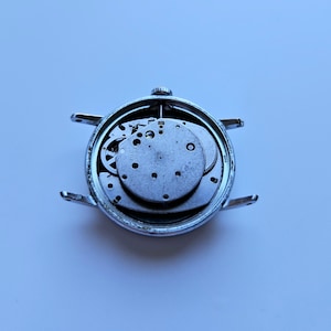 May include: A disassembled silver wristwatch, showcasing the intricate internal mechanisms. The watch face is removed, revealing gears, plates, and other components. The watch case has four lugs for attaching a strap.