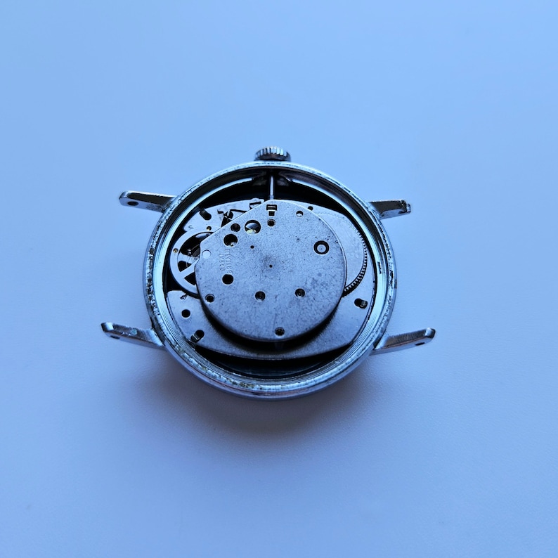 May include: A disassembled silver wristwatch, showcasing the intricate internal mechanisms. The watch face is removed, revealing gears, plates, and other components. The watch case has four lugs for attaching a strap.