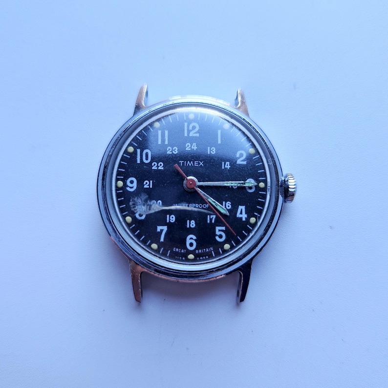 May include: A vintage Timex watch with a black face and silver casing. The watch face displays white numerals, a red second hand, and the word "TIMEX". The watch has a crown on the right side and lugs for a strap.