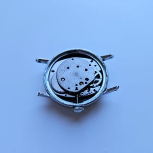 May include: A disassembled silver wristwatch with exposed internal gears and mechanisms. The watch face is missing, revealing the intricate workings of the timepiece. The watch has a small crown and lugs for attaching a strap.
