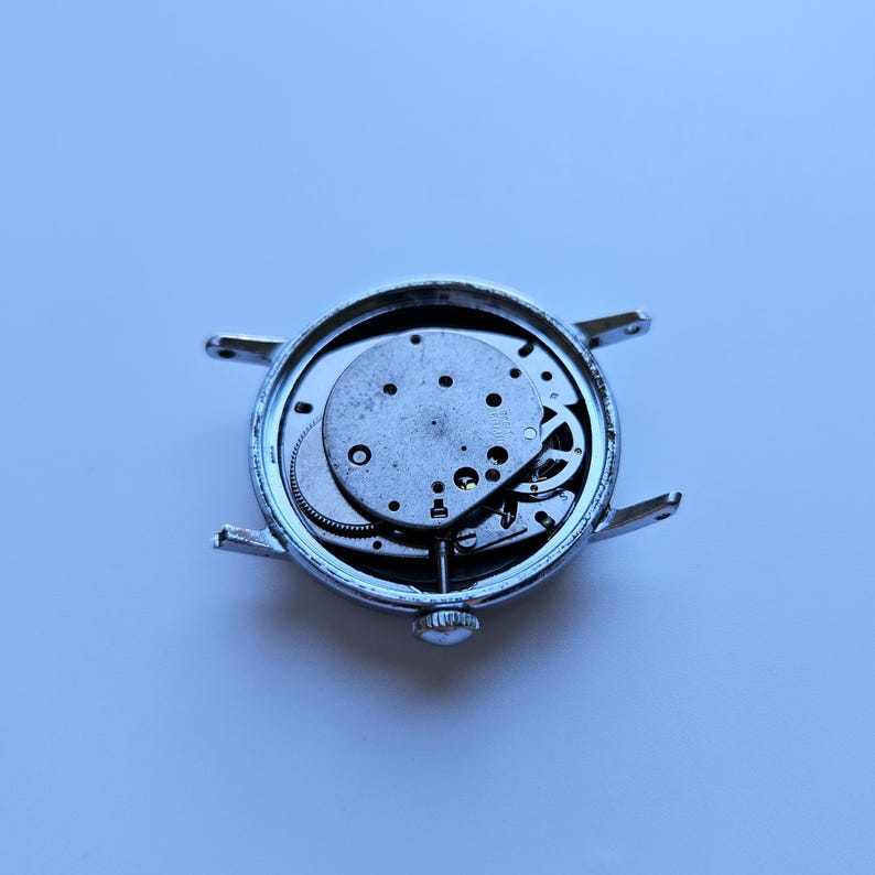 May include: A disassembled silver wristwatch with exposed internal gears and mechanisms. The watch face is missing, revealing the intricate workings of the timepiece. The watch has a small crown and lugs for attaching a strap.