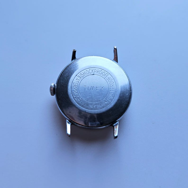 May include: A silver-toned Timex wristwatch back with the words "WATERPROOF" and "SHOCKPROOF" encircling the brand name. The watch has four lugs for attaching a strap.