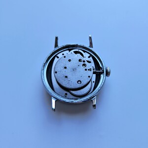 May include: A disassembled silver wristwatch with exposed internal gears and mechanisms. The watch face is missing, revealing the intricate workings of the timepiece. The watch has a silver casing and a small crown.