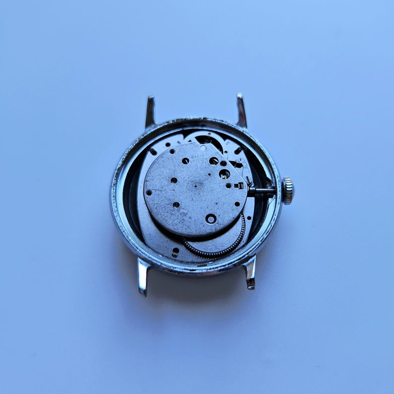 May include: A disassembled silver wristwatch with exposed internal gears and mechanisms. The watch face is missing, revealing the intricate workings of the timepiece. The watch has a silver casing and a small crown.