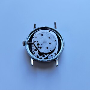 May include: A close-up view of a disassembled wristwatch, revealing the intricate internal mechanisms. The watch's silver casing surrounds the complex arrangement of gears, plates, and screws. The watch hands are missing, showcasing the inner workings of the timepiece.