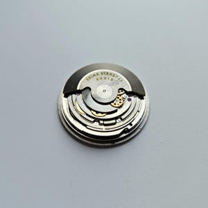 K&ouml;nnte beinhalten: Nahaufnahme eines Uhrwerks mit komplizierten Zahnr&auml;dern und Komponenten. Der Uhrenmechanismus ist kreisf&ouml;rmig, mit einer silbernen Metalloberfl&auml;che und goldfarbenen Akzenten. Der Text "BIRKS SWISS 17 JEWELS" ist sichtbar.