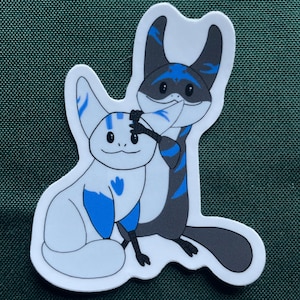May include: A sticker featuring two cartoon creatures. One is white with blue accents, the other is gray with blue stripes. They are in a playful pose, with one touching the other's head. The sticker has a white border.