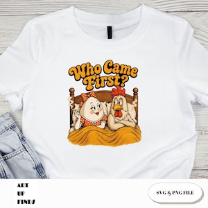 May include: White t-shirt with a humorous cartoon graphic. The graphic features an egg and a chicken in bed, with the text "Who Came First?" in orange and yellow. The egg wears a red bow and the chicken has a red comb.