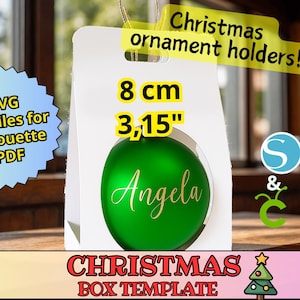 Puede incluir: Una plantilla de caja navideña blanca con una bola verde en el interior, personalizada con el nombre "Angela" en escritura dorada. La caja está etiquetada como "Christmas ornament holders!" y mide 8 cm (3,15").