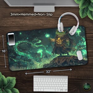 May include: A large, rectangular mousepad, 32 inches long and 16 inches wide, featuring a fantasy illustration of a frog wizard. The design includes green and blue hues with the text "3mm&bull;Hemmed&bull;Non-Slip". A smartphone, headphones, and a mouse are on the pad.