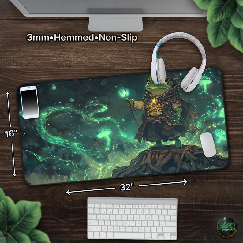 May include: A large, rectangular mousepad, 32 inches long and 16 inches wide, featuring a fantasy illustration of a frog wizard. The design includes green and blue hues with the text "3mm&bull;Hemmed&bull;Non-Slip". A smartphone, headphones, and a mouse are on the pad.
