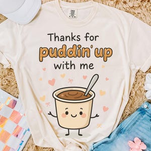 May include: Cream-colored t-shirt with the phrase "Thanks for puddin' up with me" in black and brown lettering. A cartoon pudding cup with a spoon is featured below the text, surrounded by hearts and bows.