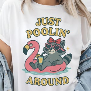 May include: White t-shirt featuring a cartoon raccoon wearing heart-shaped sunglasses and a red bow, relaxing on a pink flamingo float. The text "JUST POOLIN' AROUND" is in yellow and green.