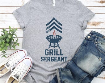 May include: A heather gray t-shirt with a distressed graphic of a barbecue grill and the words "GRILL SERGEANT" in navy blue. The shirt is paired with blue jeans and white sneakers.