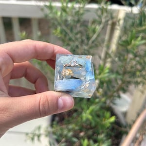 以下が含まれることがあります： 青い石の破片と金箔のフレークが入った透明な樹脂製のキューブ。上面中央には、丸い透明な石が1つ埋め込まれた銀色の指輪があり、磨き上げられた仕上げと幾何学的な形状が特徴です。