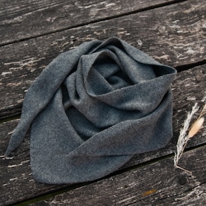 May include: A charcoal gray triangular scarf made of a soft, knit fabric. The scarf features a textured, heathered appearance with tapered ends and is arranged in loose, overlapping folds to display its flexible, draped structure.