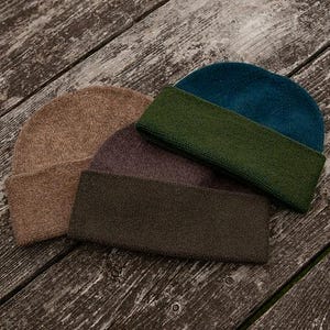 May include: A collection of four knit beanies in various colors including tan, dark brown, olive green, and teal. Each beanie features a folded cuff and a textured knit construction, displayed together in a layered arrangement.