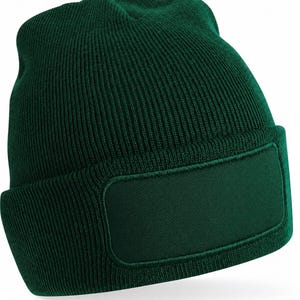 May include: A dark forest green beanie hat featuring a ribbed knit texture and a solid rectangular patch on the front cuff. The hat is constructed with a folded brim and a rounded crown, showcasing a consistent deep green colour throughout the fabric.