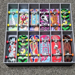 May include: A set of twelve miniature skateboards with diverse, vibrant designs, such as skulls, flames, and cartoon figures. The skateboards are displayed in a grey, compartmentalised box, each with a unique graphic.