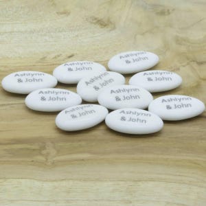Puede incluir: Un conjunto de piedras blancas de forma ovalada, cada una con los nombres "Ashlynn & John" en gris. Las piedras están dispuestas sobre una superficie de madera marrón claro, creando un artículo personalizado y decorativo.