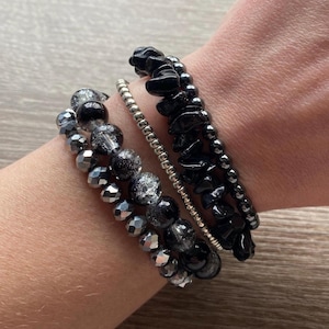 May include: A set of four bracelets featuring black and silver beads. The bracelets are stacked on a person's wrist.