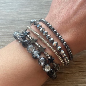May include: A set of five bracelets featuring black and silver beads with star charms. The bracelets are stacked on a person's wrist.