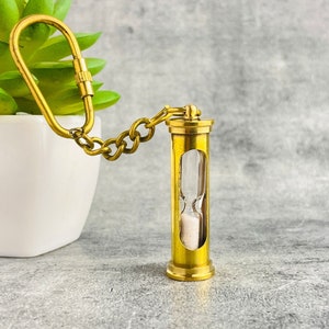 May include: A gold-toned hourglass keychain with a chain and a carabiner clip. The hourglass is filled with white sand.