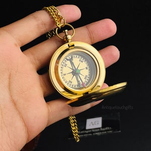 May include: A gold compass with a chain attached. The compass has a white face with black markings and a green arrow. The compass is in a hand.