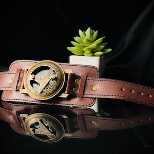 Puede incluir: Una pulsera de cuero marr&oacute;n con una cara de br&uacute;jula de lat&oacute;n. La br&uacute;jula tiene n&uacute;meros romanos y un dise&ntilde;o decorativo.