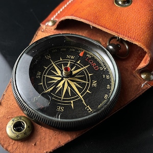 May include: A black compass with a gold needle and a gold starburst design. The compass is in a brown leather case with a white stitching detail. The compass face has the words 'LORD' and 'STEALS HUT' printed on it. The compass is pointing north.