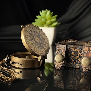 May include: A brass compass with a star design on the lid, open and revealing the compass face. The compass is attached to a chain and is sitting on a black surface next to a small, ornate, brown box.