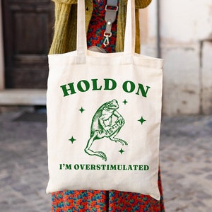 May include: A white tote bag with green text that reads "Hold On I'm Overstimulated". A green frog illustration is above the text. The bag is being held by a person.