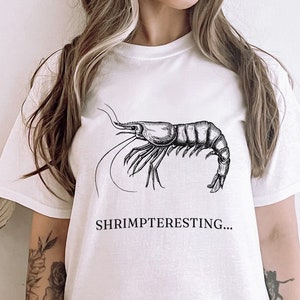 Può includere: Maglietta bianca con un'illustrazione in bianco e nero di un gambero e il testo "Shrimpteresting..."