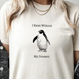 May include: A cream colored t-shirt with a black and white illustration of a penguin. The text "I Exist Without My Consent" is printed on the shirt.