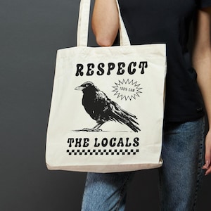 Peut inclure: Un sac fourre-tout en toile blanche avec du texte noir et une illustration de corbeau noir. Le texte dit "Respect the Locals 100% Cash".
