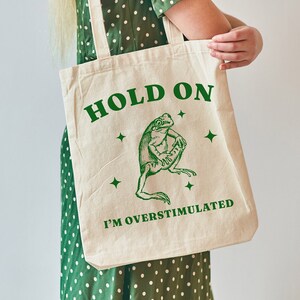 May include: A white canvas tote bag with green text that reads "Hold On I'm Overstimulated". A green frog illustration is above the text. The bag is being held by a person wearing a green polka dot dress.