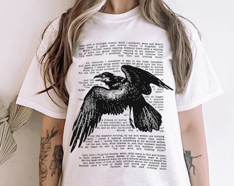 Puede incluir: Camiseta blanca con una ilustración en blanco y negro de un cuervo con el texto del poema "El Cuervo" de Edgar Allan Poe impreso en la parte delantera.