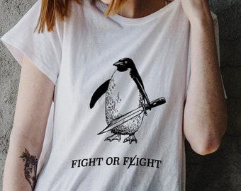 Puede incluir: Camiseta blanca con una ilustración en blanco y negro de un pingüino sosteniendo un cuchillo. El texto "Fight or Flight" está impreso debajo del pingüino.
