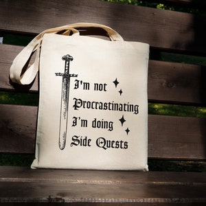 Könnte beinhalten: Eine weiße Canvas-Tragetasche mit schwarzem Text und einer Schwertdarstellung. Der Text lautet "Ich prokrastiniere nicht, ich mache Nebenquests".
