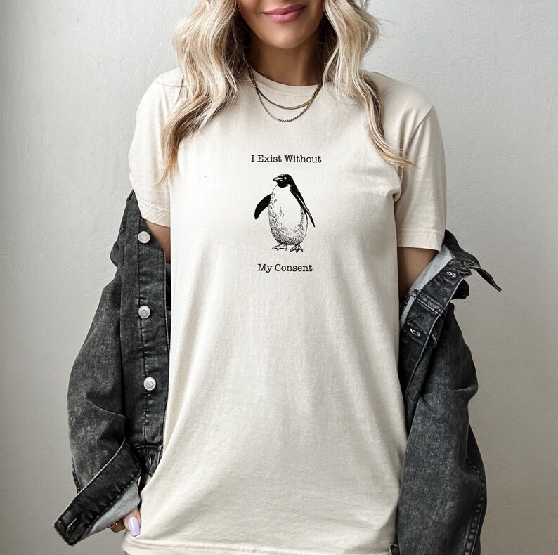 May include: A cream colored t-shirt with a black and white illustration of a penguin. The text "I Exist Without My Consent" is printed on the shirt.