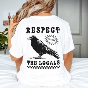 May include: White t-shirt with a black graphic of a crow and the text "RESPECT THE LOCALS." The crow is standing on a line, with the words above and below it. A starburst graphic reads "100% CAW."