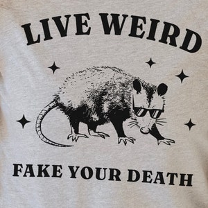 May include: Grey t-shirt with a black graphic of an opossum wearing sunglasses and the text "LIVE WEIRD FAKE YOUR DEATH".