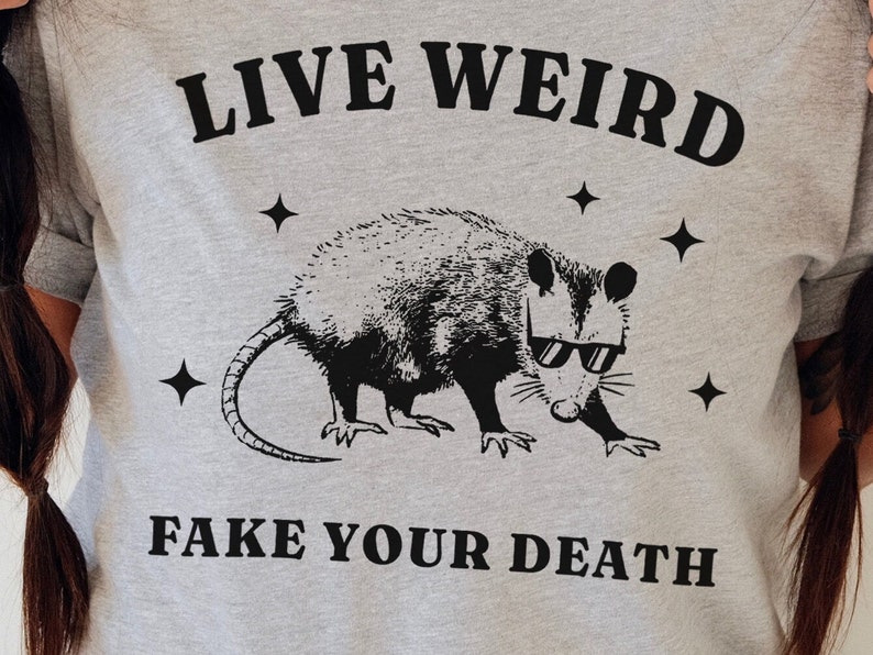 May include: Grey t-shirt with a black graphic of an opossum wearing sunglasses and the text "LIVE WEIRD FAKE YOUR DEATH".
