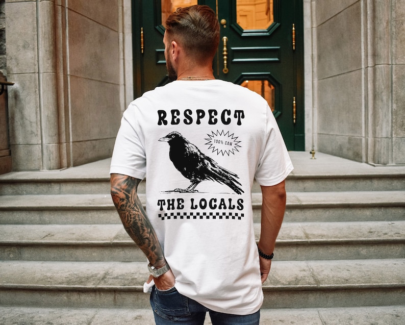 May include: White t-shirt with a black graphic of a crow and the words "RESPECT THE LOCALS". The design includes the text "100% CAW" and a checkered pattern at the bottom. The shirt is a casual wear item.