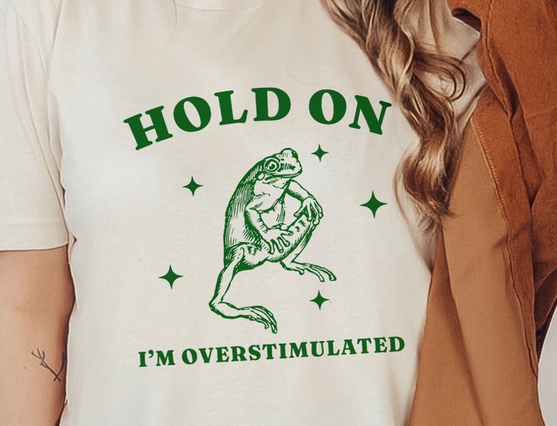 May include: White t-shirt with green text that reads "HOLD ON I'M OVERSTIMULATED". A green cartoon frog is in the center of the shirt.
