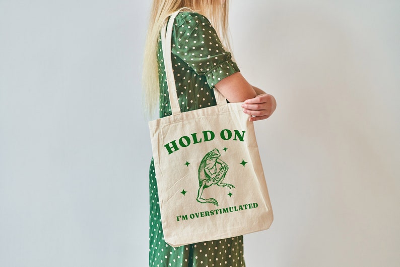 May include: A white canvas tote bag with green text that reads "Hold On I'm Overstimulated". A green frog illustration is above the text. The bag is being held by a person wearing a green polka dot dress.