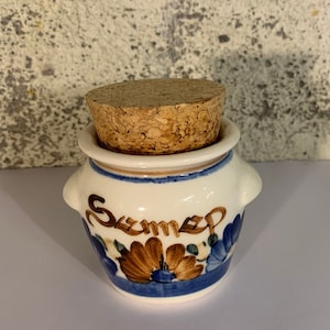 Op de afbeelding: Een kleine witte keramische pot met een kurken deksel. De pot heeft een blauw en bruin bloemenmotief en het woord "Sumez" staat op de voorkant.