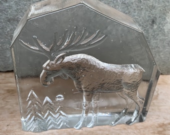 May include: A clear glass paperweight featuring an etched design of a moose standing in a snowy forest. The moose has a large set of antlers and is facing to the left. The forest is represented by a series of small, etched trees.